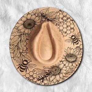 Può includere: Un cappello fedora beige con un design inciso al laser dettagliato con girasoli, api, foglie e motivi a nido d'ape. Il cappello ha una corona a goccia e un'ampia tesa.