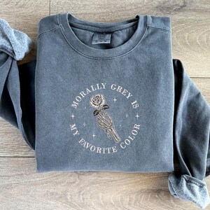May include: A gray sweatshirt with the embroidered text "MORALLY GREY IS MY FAVORITE COLOR" surrounding a skeleton hand holding a rose. The long-sleeved shirt is a muted gray color.