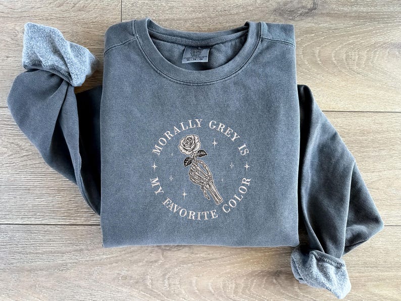 May include: A gray sweatshirt with the embroidered text "MORALLY GREY IS MY FAVORITE COLOR" surrounding a skeleton hand holding a rose. The long-sleeved shirt is a muted gray color.