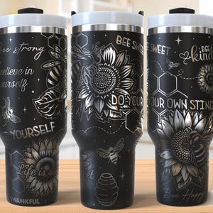 May include: Three black stainless steel tumblers with a silver design of bees, sunflowers, and honeycomb. The tumblers have different quotes on them: "Be strong believe in yourself", "Bee Sweet Do Your Own Sting", and "Bee Kind Be Happy".