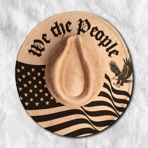 May include: Tan fedora hat with a black and white American flag design. The hat features the text "We the People" in a gothic font and an eagle graphic.
