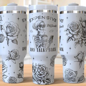 May include: White stainless steel travel mug with a black and white design featuring a skeleton holding a phone and a drink. The text on the mug reads "Expensive, Difficult and Talks Back". The mug is surrounded by black and white roses.