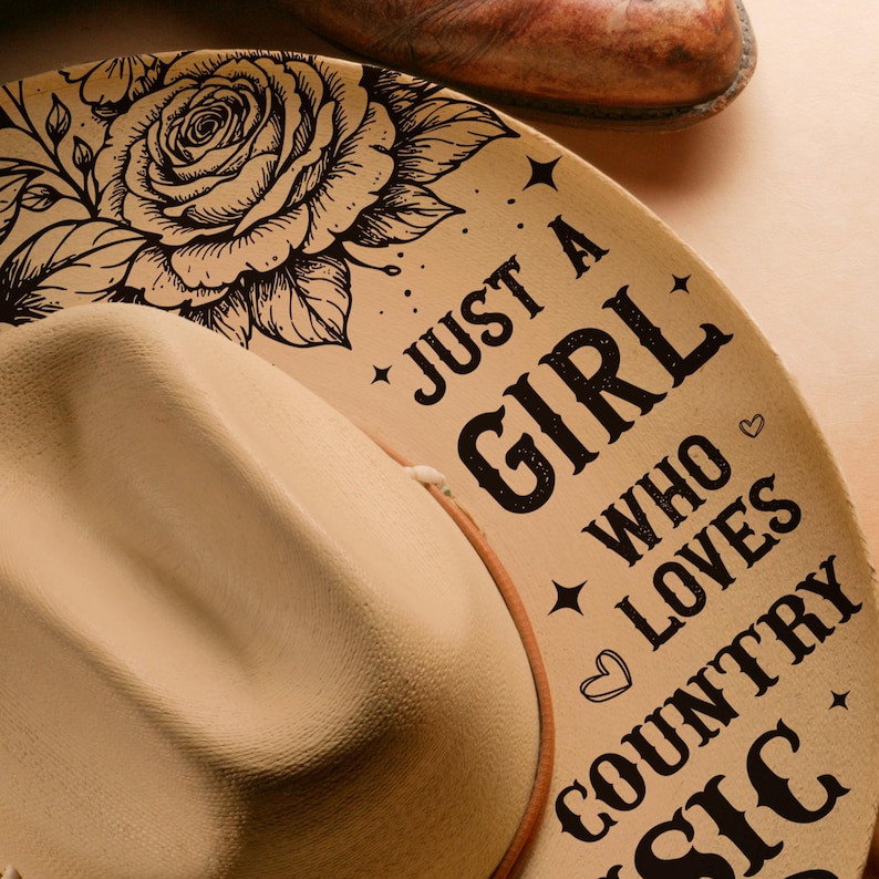 May include: A beige cowboy hat with the text "Just a Girl Who Loves Country Music" and a floral design. The hat has a brown leather band. A brown leather boot is visible in the background.