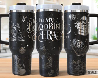 Può includere: Tazza da viaggio nera con coperchio e manico bianchi, con illustrazioni argentate di libri, fiori e un teschio. Il testo "In My Bookish Era" è visualizzato sulla tazza. Il design include stelle e dettagli floreali.