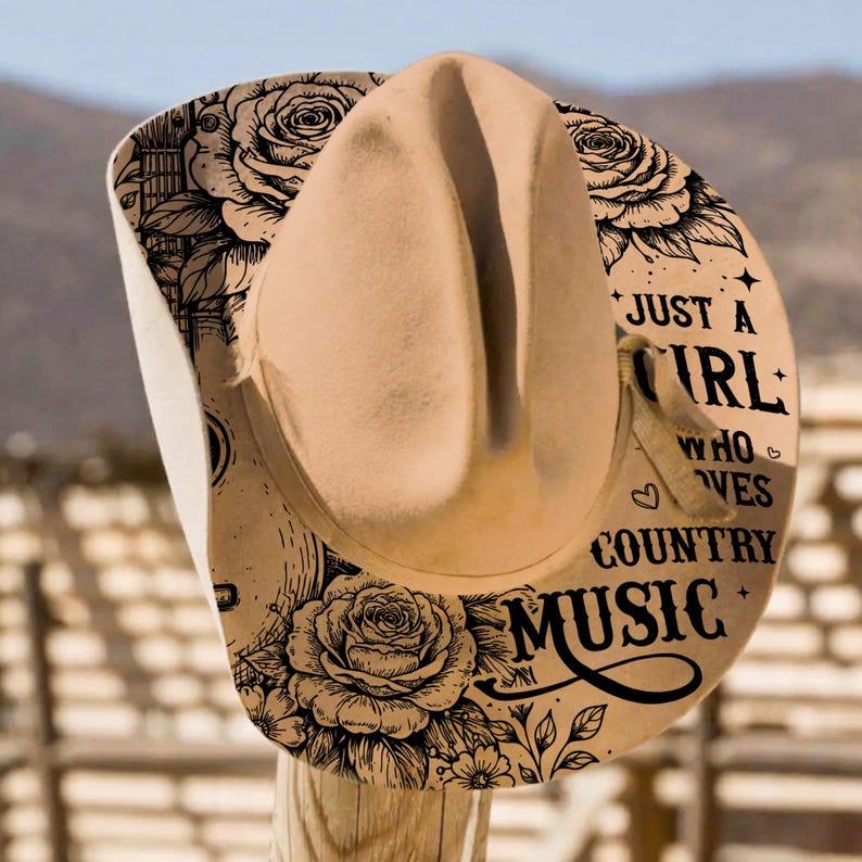 May include: A tan cowboy hat with a curved brim and a light brown crown. The hat's brim features a floral design with roses and the text "Just a Girl Who Loves Country Music."