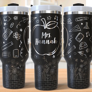 Può includere: Tazza da viaggio nera con coperchio e manico bianchi, con un design a mela argentata con il nome "Mrs. Hannah". La tazza è decorata con scarabocchi a tema scolastico e un motivo floreale alla base.