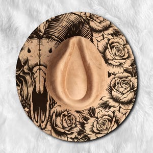 Könnte beinhalten: Ein beigefarbener Fedora-Hut mit dekorativer Krempe. Der Rand zeigt eine schwarze Grafik eines Schädels und Rosen. Der Hut hat eine tropfenförmige Krone und eine weiche, Wildleder-ähnliche Textur. Der Hut ist vor einem weißen, flauschigen Hintergrund platziert.