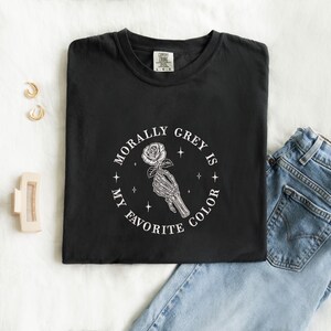 May include: Black t-shirt with the text "MORALLY GREY IS MY FAVORITE COLOR" in a circular design. The design features a skeleton hand holding a rose. The shirt is paired with blue jeans and gold earrings.