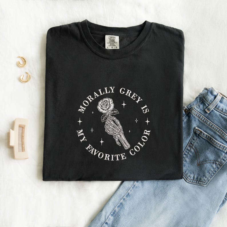 May include: Black t-shirt with the text "MORALLY GREY IS MY FAVORITE COLOR" in a circular design. The design features a skeleton hand holding a rose. The shirt is paired with blue jeans and gold earrings.