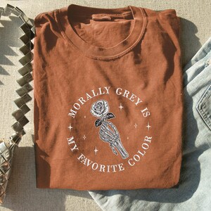May include: A rust-colored t-shirt with the text "Morally Grey Is My Favorite Color" in a circular design. The design features a skeleton hand holding a rose. The shirt is folded and placed next to a silver purse and blue jeans.