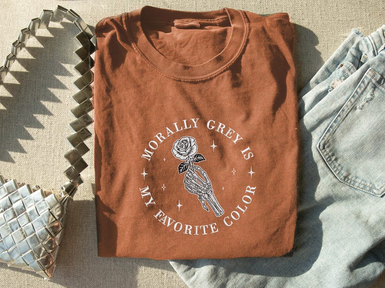 May include: A rust-colored t-shirt with the text "Morally Grey Is My Favorite Color" in a circular design. The design features a skeleton hand holding a rose. The shirt is folded and placed next to a silver purse and blue jeans.