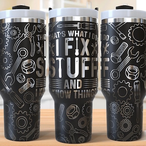 May include: Black stainless steel travel mug with a white lid and handle. The mug has a silver design of tools and the text "That's what I do I fix stuff and I know things".