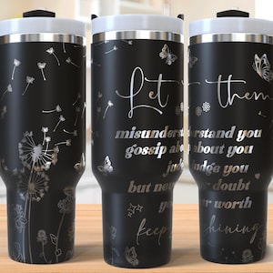 May include: Three black stainless steel travel mugs with a silver design. The mug on the left has a dandelion design. The mug in the middle has the text "Let them misunderstand you, gossip about you, judge you, but never doubt your worth. Keep shining." The mug on the right has a butterfly design.