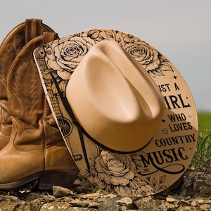 May include: A beige cowboy hat with a black band sits on a decorative board with a guitar and floral design. The board leans against brown leather cowboy boots. Text on the board says "Just a Girl Who Loves Country Music."