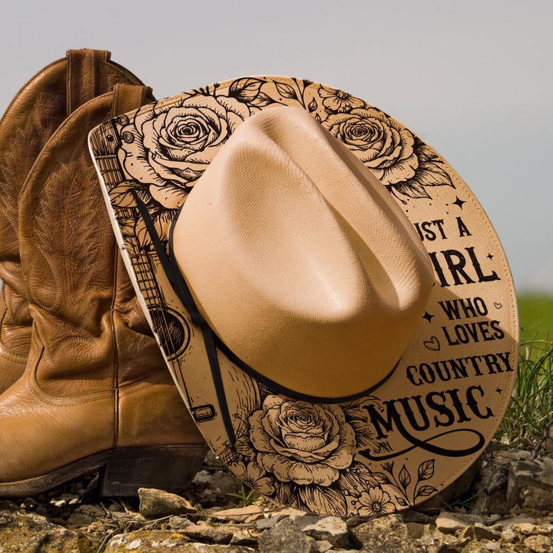 May include: A beige cowboy hat with a black band sits on a decorative board with a guitar and floral design. The board leans against brown leather cowboy boots. Text on the board says "Just a Girl Who Loves Country Music."