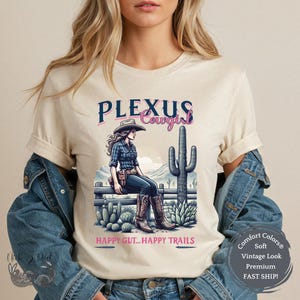 May include: A white t-shirt with a graphic design of a woman wearing a cowboy hat and sitting on a fence. The text "PLEXUS Cowgirl" is above the image and "HAPPY GUT...HAPPY TRAILS" is below the image.