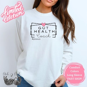 Peut inclure: T-shirt blanc à manches longues avec l'inscription "GUT HEALTH Coach" dans un design géométrique noir, un cœur rose et le hashtag #plexus. Les mots "Limited Edition" sont en script rose. Un cercle rose avec du texte est en bas à droite.