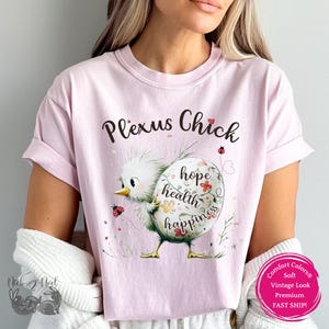 May include: A light pink t-shirt with the words "Plexus Chick" in a script font. The shirt features a cartoon chick holding an egg with the words "hope health happiness" on it. The egg is decorated with flowers and the shirt has a vintage look.