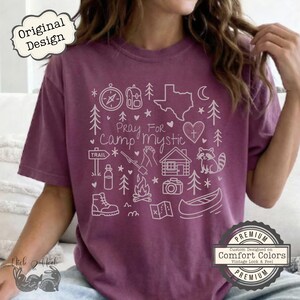 May include: A maroon t-shirt with a white graphic design featuring camping-themed illustrations and the text "Pray For Camp Mystic." The design includes a compass, backpack, Texas outline, trees, and a raccoon.