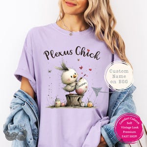 May include: A light purple t-shirt with the text "Plexus Chick" and a graphic of a chick painting an egg with the name "Susan". The design includes butterflies and hearts. The shirt has a vintage look.
