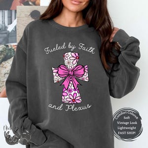 May include: A charcoal gray sweatshirt with the text "Fueled by Faith and Plexus" printed on the front. The text is in white and is centered on the sweatshirt. A pink and white floral cross is centered above the text. The sweatshirt has a distressed look and is made of a soft, comfortable fabric.