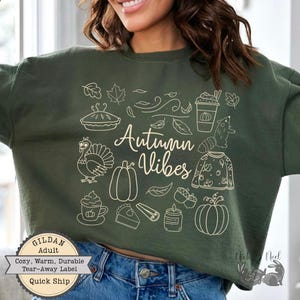 May include: Dark green sweatshirt featuring the cursive text "Autumn Vibes" and autumn-themed illustrations. The design includes a pumpkin, turkey, pie, and leaves. The sweatshirt has a "GILDAN Adult" label.