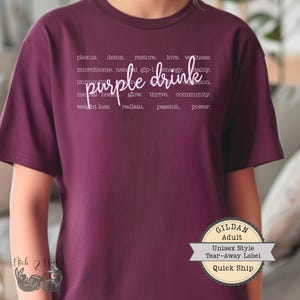 May include: A maroon unisex t-shirt with the words "purple drink" in a cursive font. Surrounding the text are words like "detox," "restore," and "vitality." The shirt has a tear-away label and is labeled "Quick Ship."