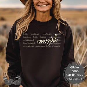 May include: Black sweatshirt with the word "cowgirl" in a cursive font. The sweatshirt also has a list of words like "bold," "fearless," and "courageous." The Gildan 18000 sweatshirt is soft and unisex.