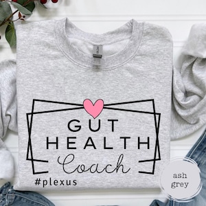 May include: A light gray sweatshirt with a black and white graphic design that says "Gut Health Coach" with a pink heart above the text and the hashtag "#plexus" below. The sweatshirt is on a white surface with a blue denim background.