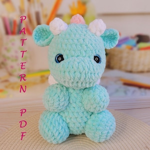 May include: A handmade, light blue crochet giraffe with white and pink accents. The plush toy has large blue eyes and is sitting upright. The word "PATTERN PDF" is written vertically in pink letters on the left side.