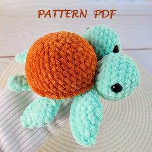 May include: A crocheted turtle toy, featuring an orange shell and teal head and flippers. The image includes the text "PATTERN PDF". The turtle is made with a textured yarn and rests on a white surface. The toy is a handmade item.