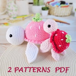 Puede incluir: Peluche de tortuga de fresa de ganchillo en rosa y rojo con detalles blancos y tallos verdes. La tortuga tiene una cabeza y aletas blancas. El texto "2 PATTERNS PDF" se muestra en la parte inferior.