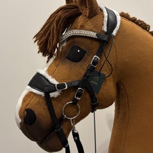 Puede incluir: Una cabeza de caballo de peluche marrón con una brida negra y una banda frontal plateada. La brida tiene correas de cuero negro y hebillas plateadas. El caballo tiene una melena marrón y una etiqueta que dice "Off Tack".