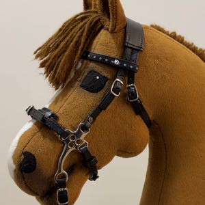 May include: A plush brown hobby horse head with a black leather bridle. The bridle features silver-colored metal hardware and decorative rhinestones. The horse has a white stripe on its face and a brown mane.