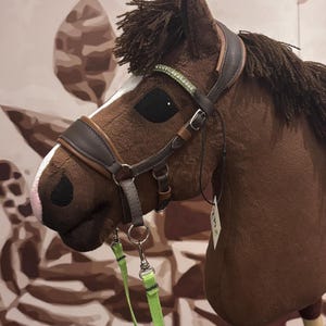 May include: A brown hobby horse with a detailed leather bridle and green reins. The horse has a white stripe on its face and a decorative band with green accents on the bridle. The mane and forelock are made of textured brown material.