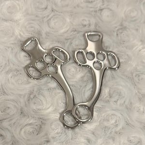 May include: Two polished silver horse bits, each with a series of holes and rounded edges. The bits have a curved design and a ring at the bottom for reins. The background is a soft, white, textured fabric.