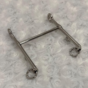 Könnte beinhalten: Ein silberfarbenes Metallgebiss mit einem geraden Steg und zwei abgewinkelten Seitenteilen, jeweils mit einem kleinen Ring. Das Gebiss ist für die Verwendung mit dem Maul und den Zügeln eines Pferdes konzipiert.