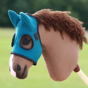 May include: A plush toy horse head with a teal mask and brown mane. The horse has a light brown body and a wooden stick handle. The toy is designed for children's play and imaginative games.