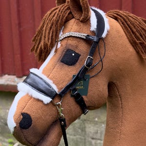 May include: A brown plush hobby horse with a white blaze on its face and brown yarn mane. It wears a black leather bridle featuring white faux fur padding, a browband with clear rhinestones, and a small dark green tag labelled OR Tack.