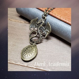 May include: A bronze dragon pendant necklace with a circular locket. The locket has an intricate design with a floral pattern. The necklace is on a brown leather surface with the text "Dark Academia" visible.