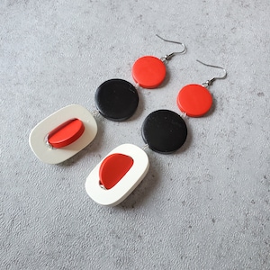 May include: A pair of modern earrings featuring a geometric design. Each earring has a white, oval-shaped element with a red insert, connected to a black disc and a red disc. The earrings hang from silver-colored hooks.