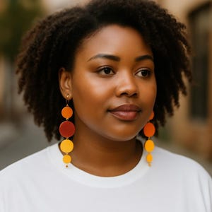 May include: A woman wearing long, dangling earrings with a gradient of colors. The earrings feature circular shapes in shades of yellow, orange, and red. The woman is wearing a white t-shirt.