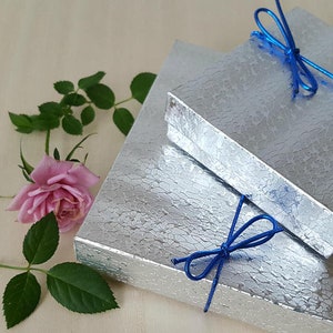 May include: Two silver gift boxes with blue ribbon bows. The boxes are stacked on top of each other. The top box has a blue ribbon tied in a bow on top. The bottom box has a blue ribbon tied in a bow on the side. The boxes are on a light wood surface.