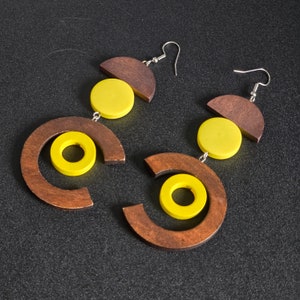 May include: A pair of geometric earrings featuring a combination of dark brown wood and bright yellow circular accents. The earrings have a unique design with a semi-circle, a circle, and a smaller circle, connected by silver-colored hooks.