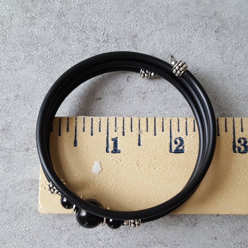 May include: Black rubber bracelet with silver accents and black beads. The bracelet is measured against a ruler, showing it is approximately 11 inches in circumference.
