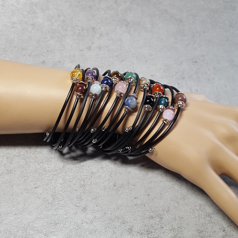 May include: A stack of black elastic bracelets with colorful gemstone beads. The bracelets are layered on a person's wrist.