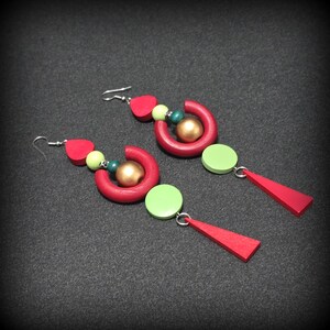 May include: A pair of handmade earrings with a bold, geometric design. The earrings feature red, green, and gold elements, including a red half-circle, a green circle, and a red triangle. The earrings are suspended from silver-colored hooks.
