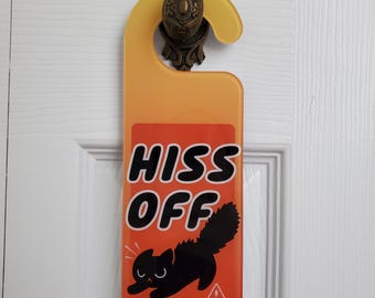 Op de afbeelding: Een geel en oranje deurhanger met een illustratie van een zwarte kat en de tekst HISS OFF en [RIGHT MEOW] in zwarte letters. Het ontwerp bevat een klein wit waarschuwingsdriehoekje met een uitroepteken op een rechthoekige achtergrond.