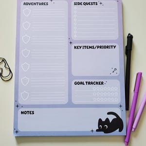 May include: A notepad with sections for "Adventures," "Side Quests," "Key Items/Priority," and "Goal Tracker." The notepad has a gradient blue and purple background with a black cat illustration. A black pen and two purple pens are next to the notepad.