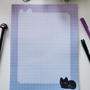 May include: A notepad with a gradient blue and purple background and a grid pattern. The top of the notepad has a cat ear design, and a black cat illustration is at the bottom. A black pen with a paw print and a black cat pen are on the sides.